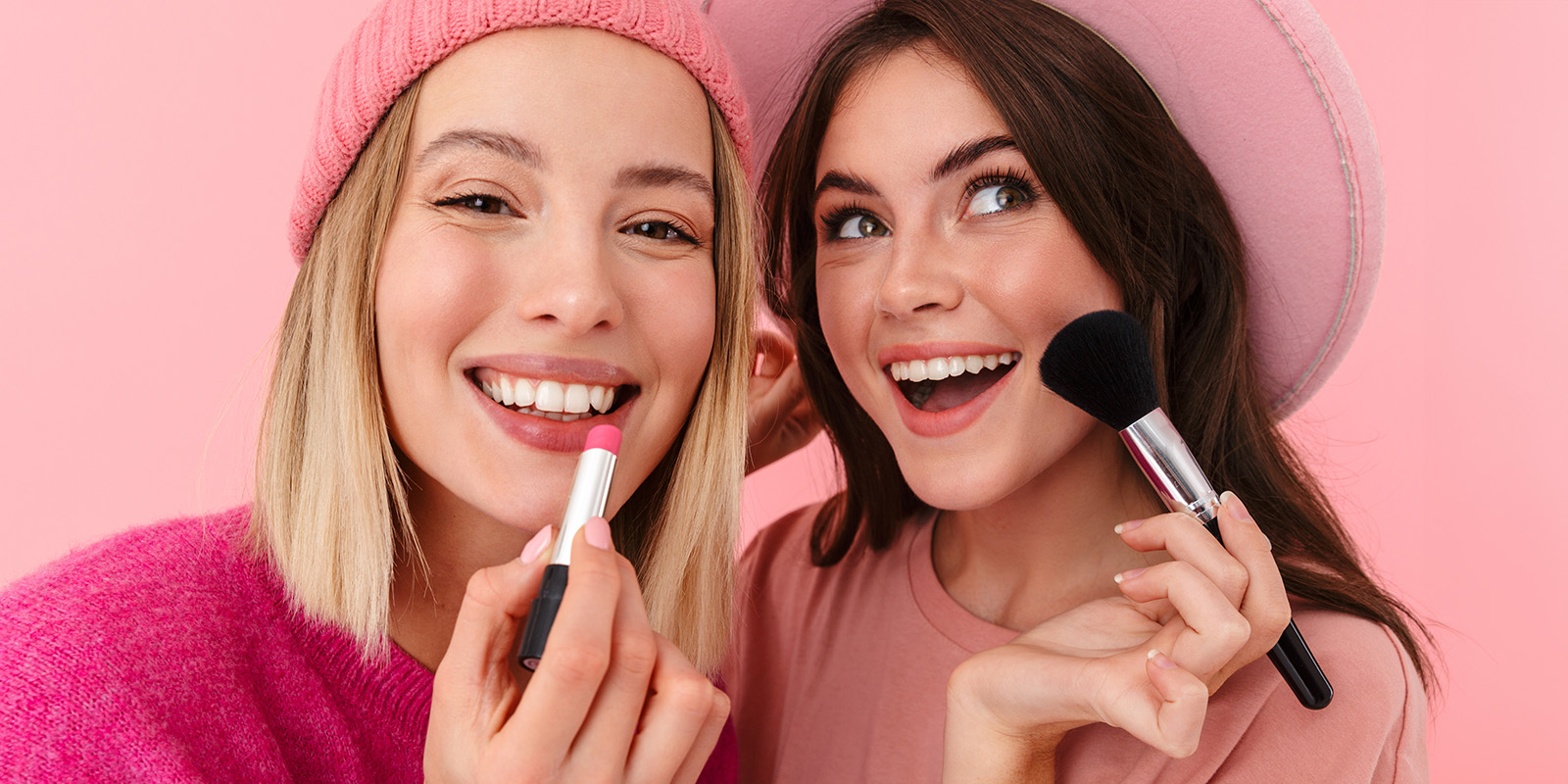 Zwei lachende Frauen, die ien hat einen Lippenstift in der Hand, die andere eine Kosmetikpinsel.