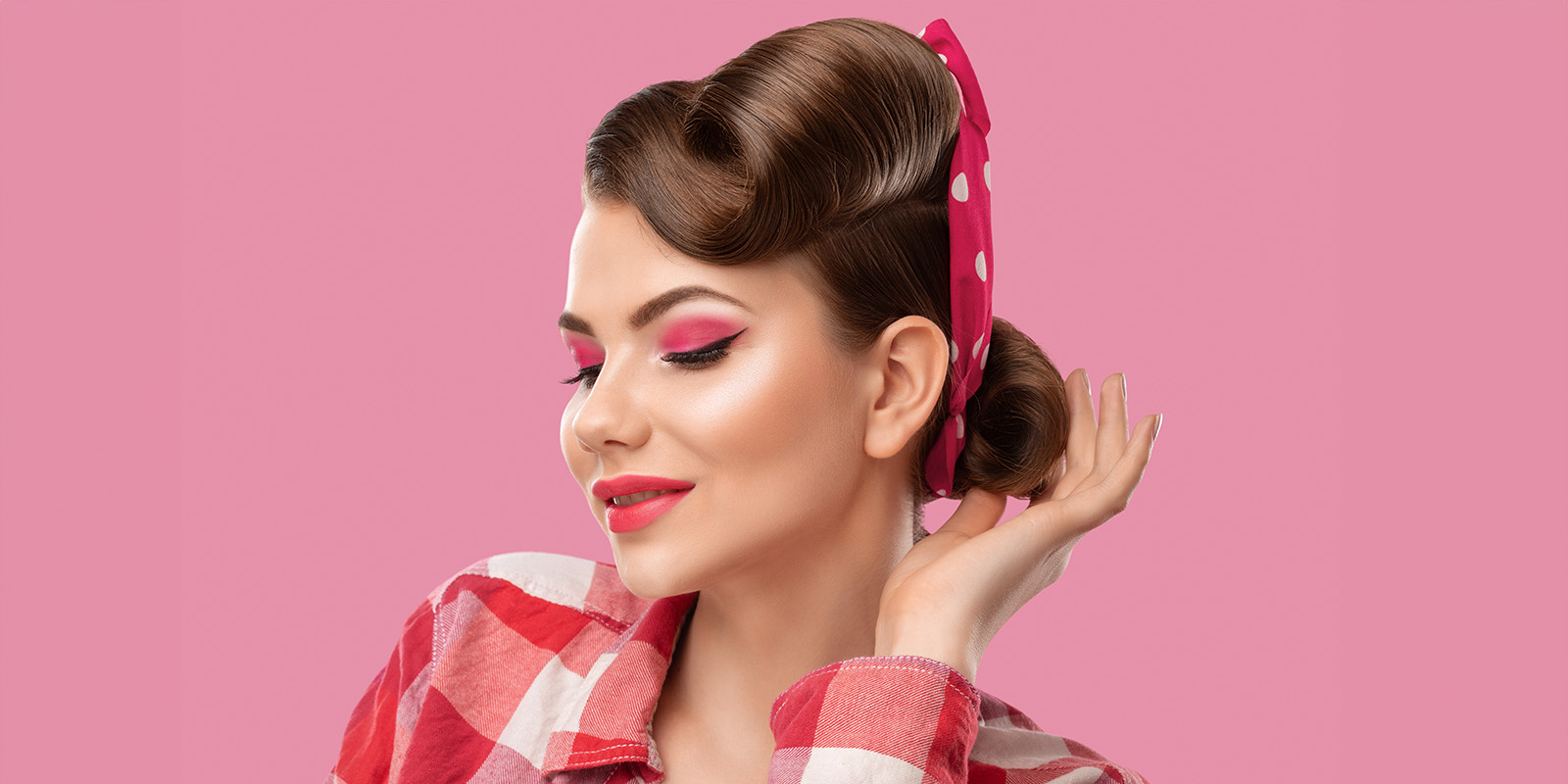 Eine hübsche Frau mit Make-Up und Haaren im Stil der 50er-Jahre schaut leicht nach unter und greift mit der Hand an die Frisur.