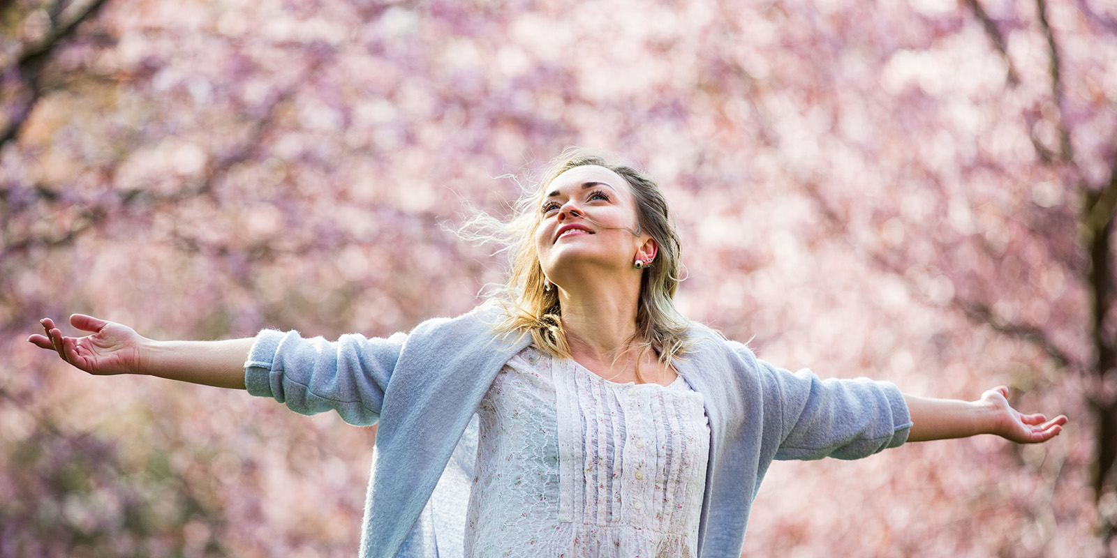 Blonde Frau schaut zum Himmel und breitet Arme aus vor rosa blühenden Bäumen im Hintergrund.