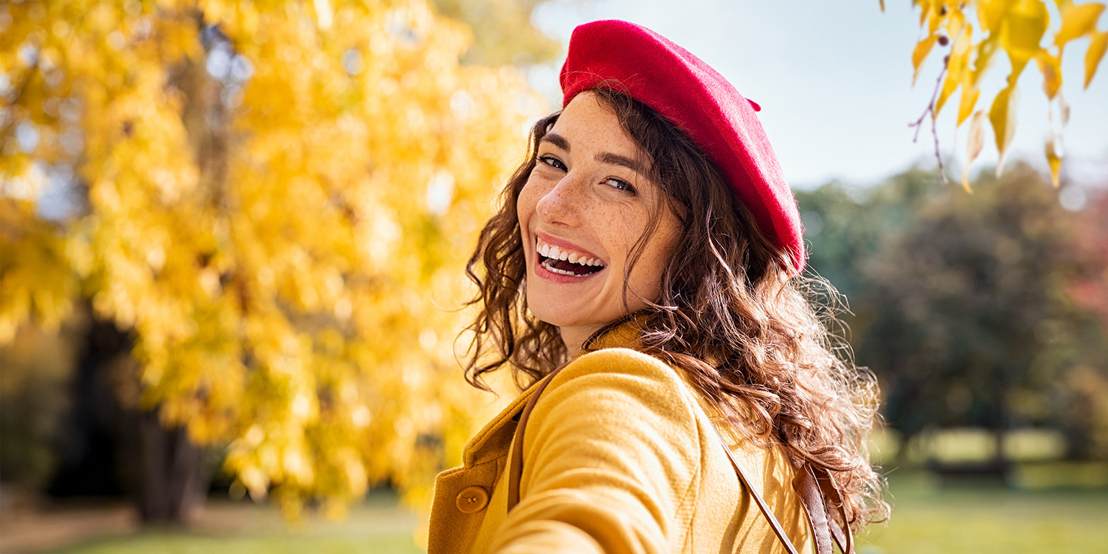 frau-herbst-gesicht EIne Frau mit langen gelockten Haaren und Baskenmütze lacht in die Kamera vor einem herbstlichen Hintergrund.