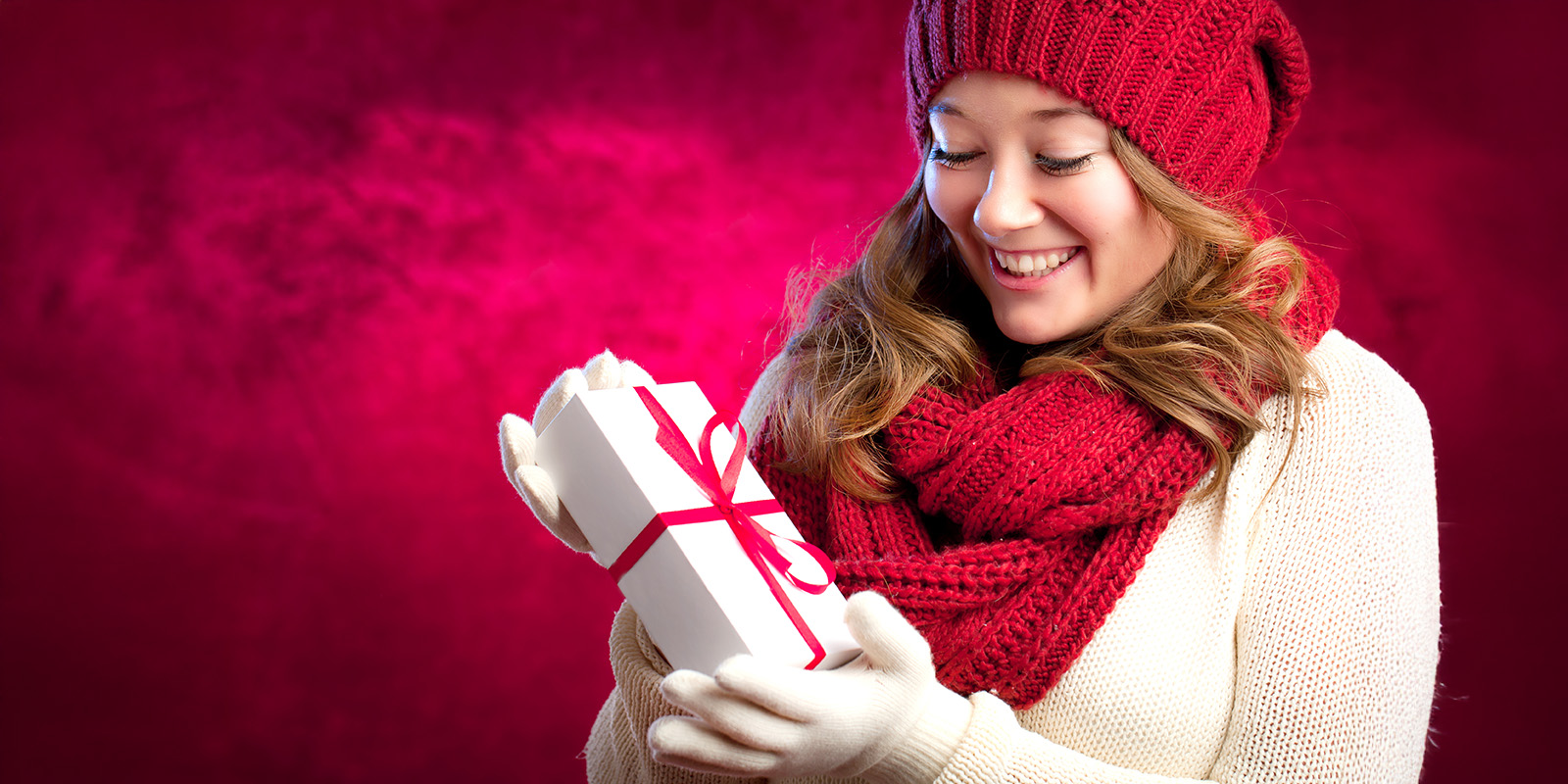 Eine lachende junge Frau mit Mütze und Handschuhen hält vor einem roten Hintergrund ein Geschenkpäckchen in den Händen.