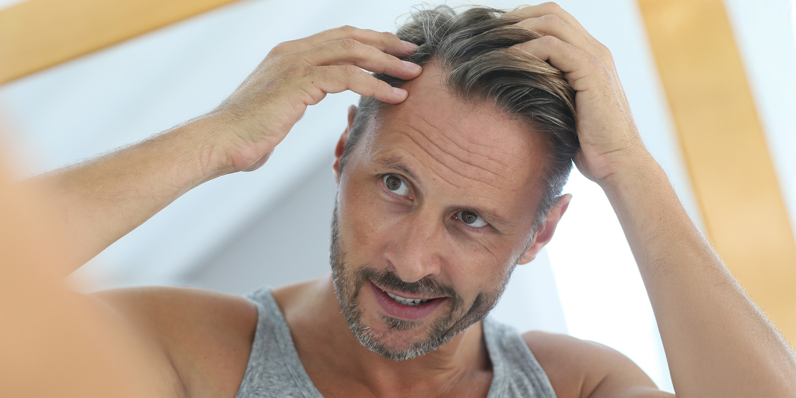 Ein Mann mit angegrauten Haaren steht vor einem Spiegel und untersucht mit beiden Händen seinen Haaransatz auf Haarausfall und grauem Haar.