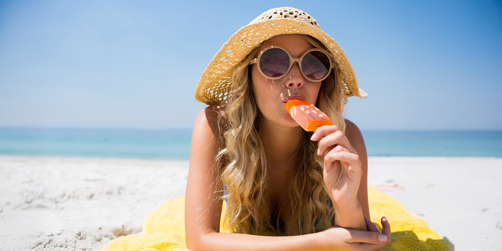 Eine Frau mit Hut und Sonnenbrille liegt, auf ihre Arme gestützt, in der Sonne am Strand und isst ein Eis am Stiel.