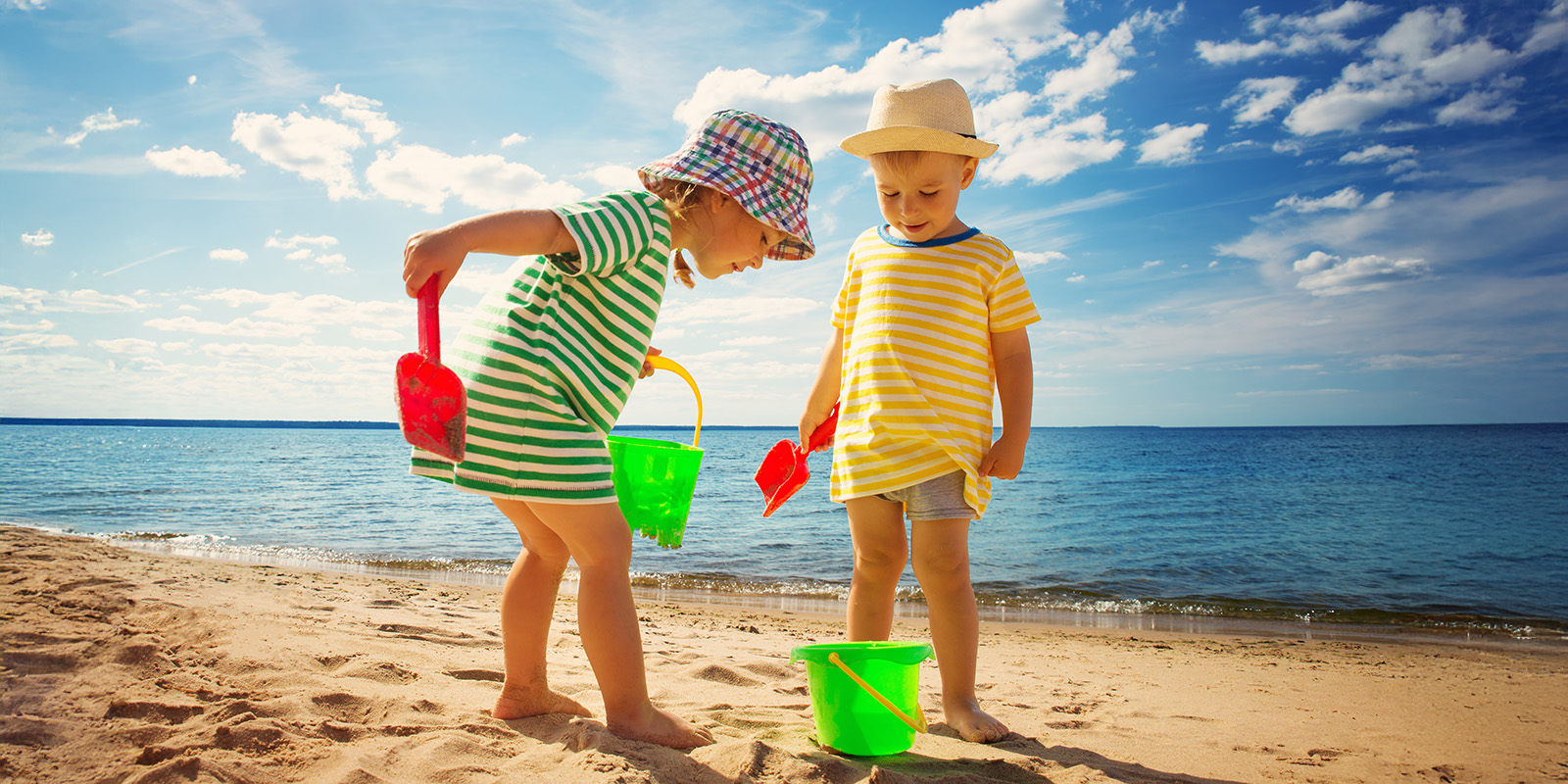 Zwei Kinder mit Bekleidung und Sonnenhut spielen mit Eimer und Schaufel am Strand.