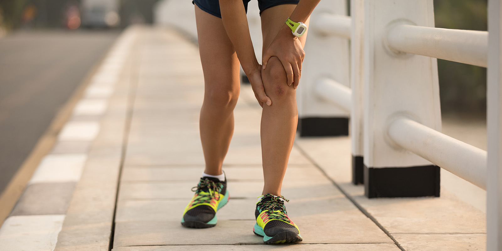 Eine Person in sportlicher Kleidung und Joggingschuhen fasst sich mit ihren Händen an das linke Knie.