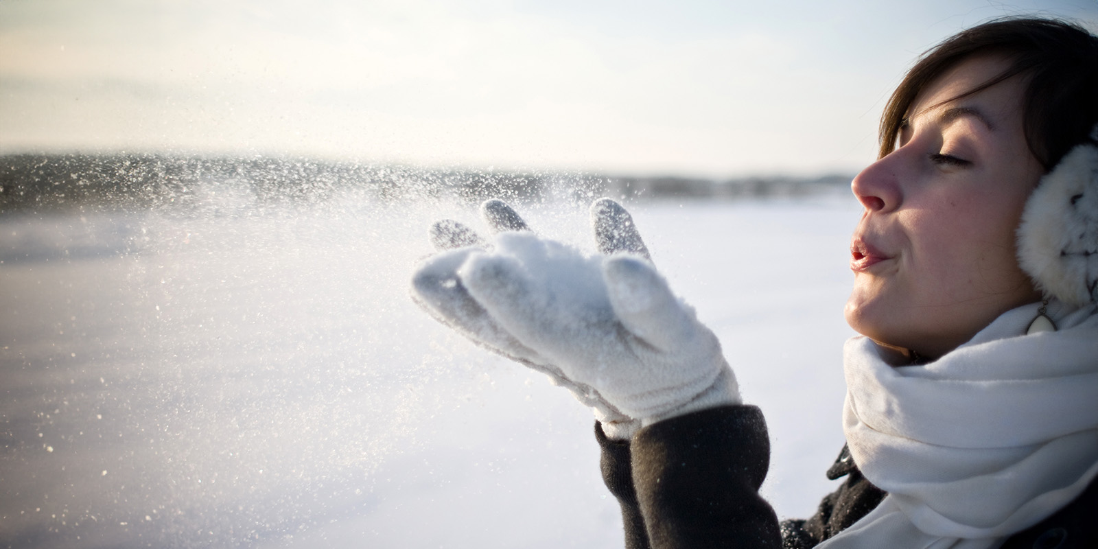 Eine Frau hält etwas Schnee mit Handschuhen an den Händen hoch und pustet ihn in die Luft.