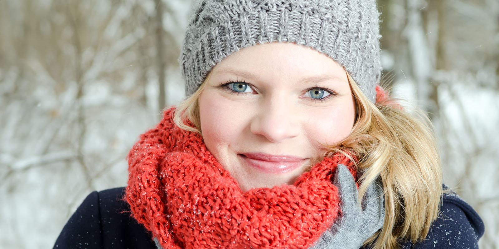 Eine hübsche Frau mit Mütze und Schal schaut lächelnd in die Kamera - im Hintergrund sieht man einen winterlichen Wald.